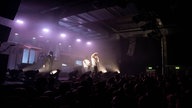 Blick auf eine lila beleuchtete Bühne, auf der drei Musiker zu sehen sind, in einer dunklen Veranstaltungshalle voll mit Menschen. 