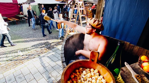 Ein Mann sitzt oberkörperfrei in einer Badewanne aus Holz. Hinter ihm laufen Besucher den Weg entlang.