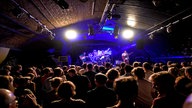 Innenraum eines Clubs, mehrere Menschen schauen auf eine blau beleuchtete Bühne, auf der eine Band steht.