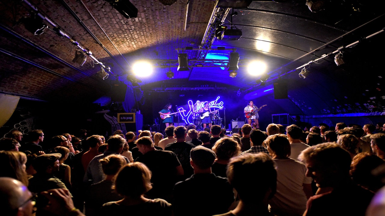 Innenraum eines Clubs, mehrere Menschen schauen auf eine blau beleuchtete Bühne, auf der eine Band steht.