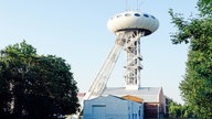 Ein schmaler Turm ragt aus einem Gebäude heraus. Oben auf dem Turm sitzt ein Ufo-ähnliches Gebilde.