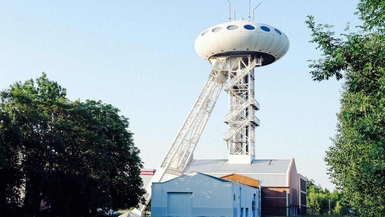 Ein schmaler Turm ragt aus einem Gebäude heraus. Oben auf dem Turm sitzt ein Ufo-ähnliches Gebilde.