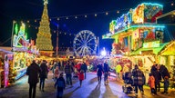 Menschen gehen zwischen bunt leuchtenden Weihnachtsmarktständen und Kirmesbuden. Im Hintergrund blaues Riesenrad und großer Tannenbaum.
