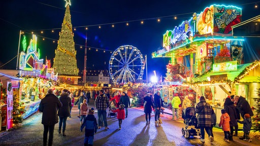 Menschen gehen zwischen bunt leuchtenden Weihnachtsmarktständen und Kirmesbuden. Im Hintergrund blaues Riesenrad und großer Tannenbaum.