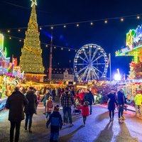 Menschen gehen zwischen bunt leuchtenden Weihnachtsmarktständen und Kirmesbuden. Im Hintergrund blaues Riesenrad und großer Tannenbaum.