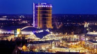 Ein riesiges, bunt beleuchtetes Industriedenkmal ragt über eine moderne Stadtkulisse, in der hell erleuchtete Gebäude, Straßen und das umliegende Stadtgebiet glitzern.