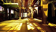 Das Innere der alten Industrieanlage mit vielen Stahlträgern, Rohren und Gerüsten, durch die warmes gelbliches Licht fällt.