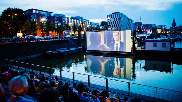 Eine Szene des Open-Air-Kinos am Kölner Rheinauhafen, mit dem Hafen im Vordergrund und dem Kinoareal im Hintergrund.