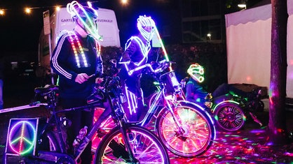 Zwei Männer mit Fahrrädern stehen nebeneinander. Es ist dunkel, doch sie leuchten bunt, weil sie mit vielen LED-Lichtern ausgestattet sind.