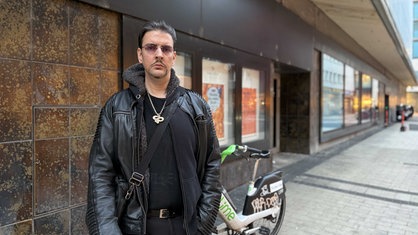 Markus R., komplett in Schwarz gekleidet, mit goldener Schlangen-Kette um den Hals, eine Umhängetasche über der Schulter, steht vor einem Geschäft in der Kölner Innenstadt