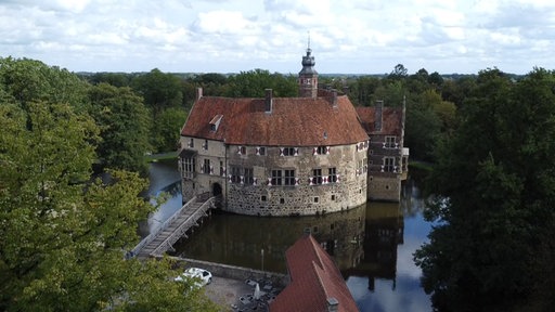 Blick auf eine alte Wasserburg aus Stein mit rotem Ziegeldach.
