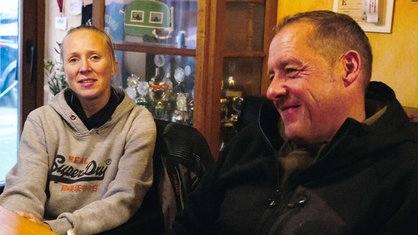 Eva Wissing, eine blonde Frau im grauen Pullover, sitzt vor einer Glasvitrine mit Pokalen. Rechts neben ihr sitzt ihr Mann, Dirk Wissing.
