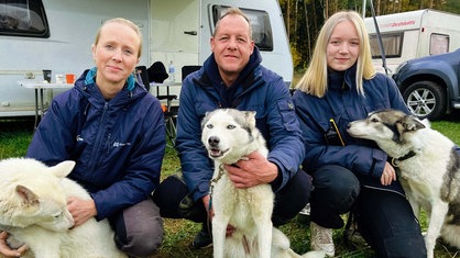 Mutter Eva, Vater Dirk und Tochter Aaliyah Wissing, hocken vor einem grauen Wohnwagen und halten drei Huskys in ihren Armen.