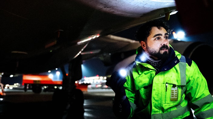Fluggerätmechaniker Adam kontrolliert die Außenwand eines Flugzeugs, in der Hand hält er eine Taschenlampe.