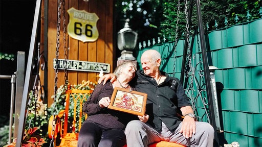 Fred und Ed Asbeck sitzen auf einer Schaukel vor einer grünen Wand und einer Holzpanele, an der ein Route-66-Schild hängt.