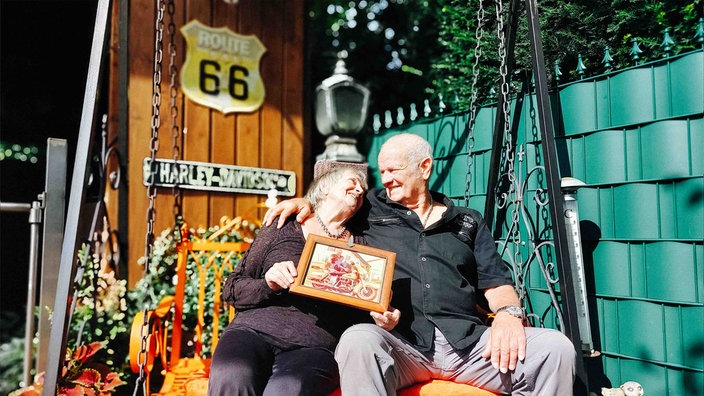 Fred und Ed Asbeck sitzen auf einer Schaukel vor einer grünen Wand und einer Holzpanele, an der ein Route-66-Schild hängt.