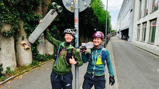 Zwei Frauen stehen auf einer Straße, sie tragen Helme und haben jeweils ein Longboard in der Hand