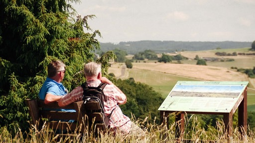 Ausblick vom Kalvarienberg auf dem Wanderweg "Eifelspur Toskana". Zwei Männer sitzen auf einer Bank und schauen in die Ferne.