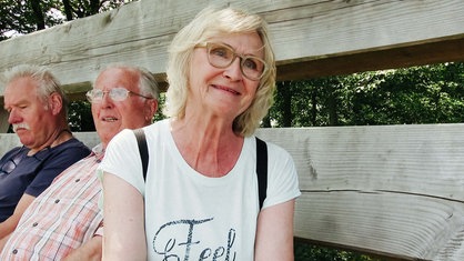 Monika Bolder sitzt auf einer großen Holzbank. Links neben ihr sitzen zwei Männer. Sie lächelt.