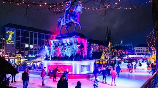 eine Eisbahn mit bunter Beleuchtung, die um ein Reiterdenkmal führt, im Hintergrund ist der Kölner Dom zu sehen