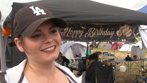 Frau mit schwarzer Cap, Haare hinten zusammengebunden, lächelt, im Hintergrund sieht man einen Zeltplatz, auf einer Plane steht "Happy Birthday".
