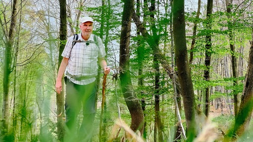 Ralf Spilles, ein Mann mit Kappe und leichtem weißem Bart und Gehstock in der Hand. Im Hintergrund ein dicht bewachsenener Wald.