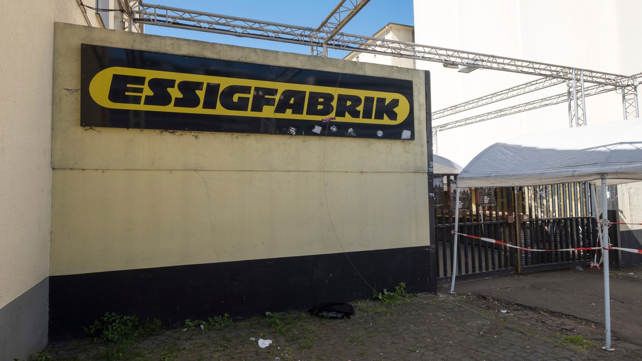 Ein beige-farbenes, verputztes Gebäude mit einem großen, schwarz-gelben Schriftzug, auf dem Essigfabrik drauf steht. Über dem Gebäude sind Traversen aufgebaut, rechts ist der Eingang mit Absperrband und einem Tor abgesperrt.