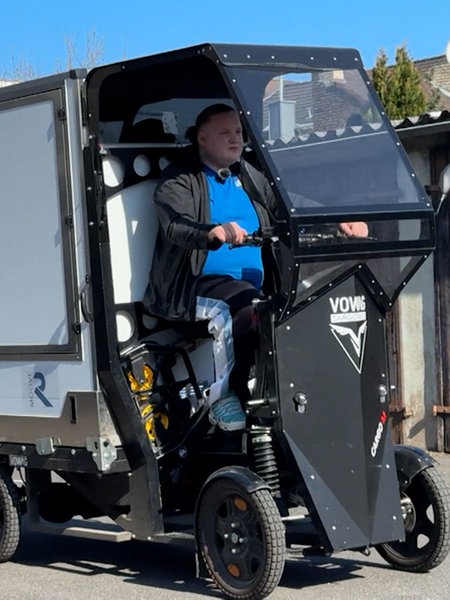 Fabian Klinz fährt ein umgebautes elektrisches Lastenrad mit Bedachung, Transportkasten und Anhänger auf einem Betriebsgelände. Im Hintergrund Garagentore