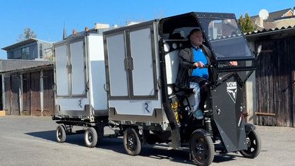 Fabian Klinz fährt ein umgebautes elektrisches Lastenrad mit Bedachung, Transportkasten und Anhänger auf einem Betriebsgelände. Im Hintergrund Garagentore