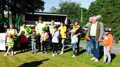 Mehrere Erwachsene und Kinder mit Helm und gelber Warnweste stehen für ein Gruppenfoto auf einer Wiese.