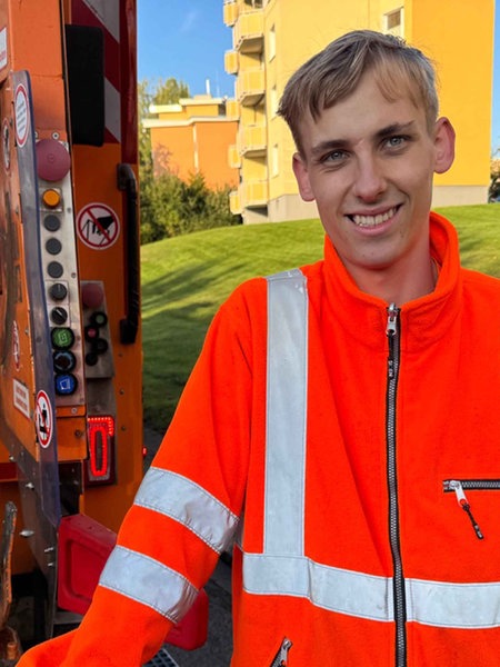 Laurin Oppermann steht im orangenen Overall vor einem Müllfahrzeug.