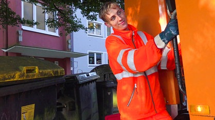 Laurin Oppermann steht mit orangenem Overall auf einem Müllfahrzeug. 