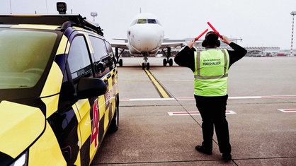 David Schönemann steht neben dem Follow-Me-Car auf dem Vorfeld und winkt ein Flugzeug ein.