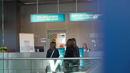 Der Schalter der Flughafen-Seelsorge in Düsseldorf, dahinter zwei Ehrenamtliche, davor Passagiere