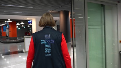 Seelsorgerin Ute Clevers läuft durch den Düsseldorfer Flughafen, auf dem Rücken ihrer Weste stehen das Wort "Willkommen" in verschiedenen Sprachen