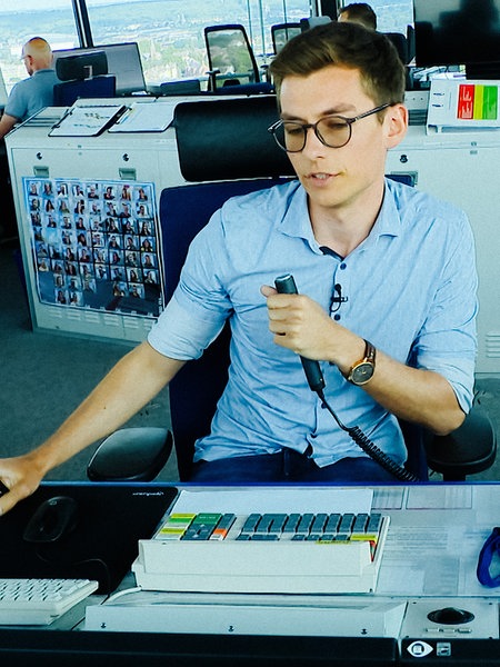 Jannik Ostendorf sitzt im Flughafen-Tower an einem Schreibtisch, hat ein Mikrofon in der Hand und schaut dabei auf Bildschirme. 