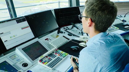 Schulterblick auf den Arbeitsplatz von Fluglotse Jannik Ostendorf. Drei Bildschirme, zwei Mäuse und mehrere Mikrofone sind vor ihm.