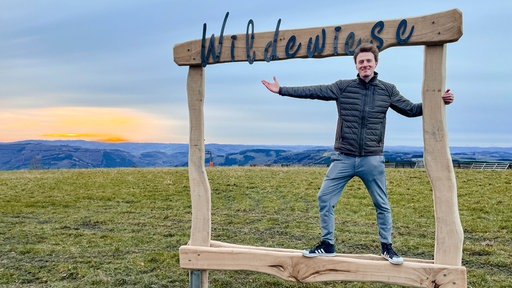 Ein Holzrahmen in der Natur des kleinen Ortes Wildewiese. Darin steht ein Mann und lächelt in die Kamera.