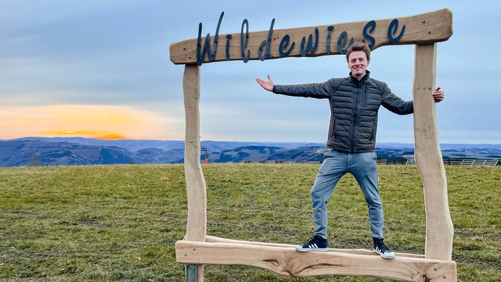 Ein Holzrahmen in der Natur des kleinen Ortes Wildewiese. Darin steht ein Mann und lächelt in die Kamera.