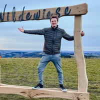 Ein Holzrahmen in der Natur des kleinen Ortes Wildewiese. Darin steht ein Mann und lächelt in die Kamera.