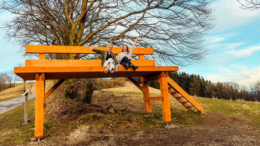 Eine XXL-Holzbank im kleinen Ort Wildewiese. Zwei Frauen sitzen auf der Bank, strecken ihre Arme aus und lächeln in die Kamera.
