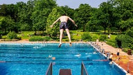 Junge springt vornüber von einem 3-Meter-Brett, steht in der Luft, unter ihm der Pool mit einigen Menschen im Pool und am Beckenrand, in naher Ferne Liegewiese und Wald 