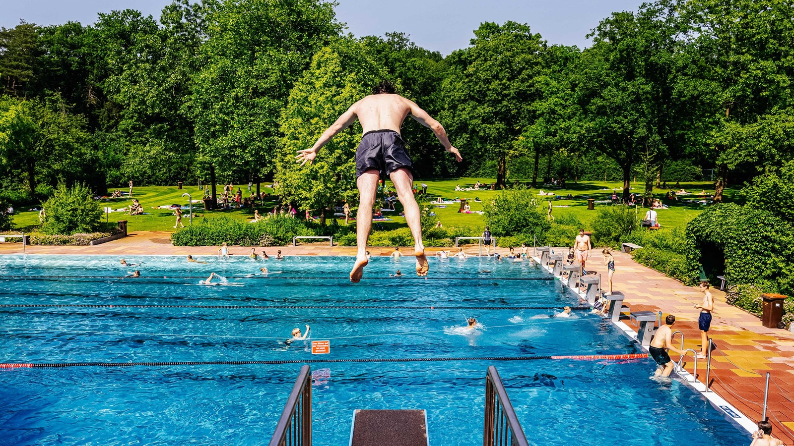 Junge springt vornüber von einem 3-Meter-Brett, steht in der Luft, unter ihm der Pool mit einigen Menschen im Pool und am Beckenrand, in naher Ferne Liegewiese und Wald 