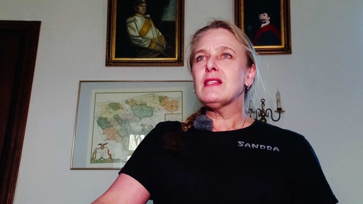 Eine Frau mit blonden Haaren und schwarzem T-Shirt. Auf dem Shirt steht rechts auf der Brust in weißer Schrift "Sandra".