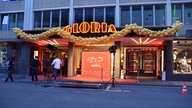 Ein Gebäude von außen, geprägt von einem großen, rot-gelben Schriftzug auf dem Gloria steht. Darunter ein großer Eingang mit rotem Teppich und vielen goldenen Ballons.