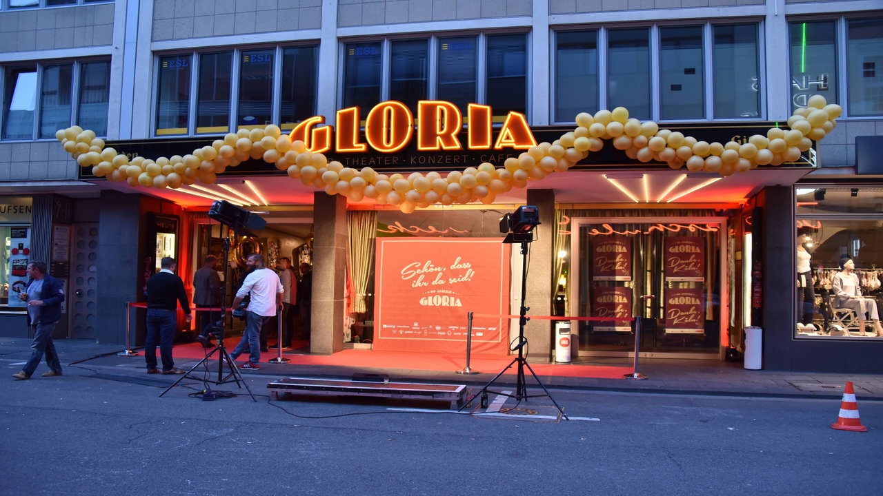 Ein Gebäude von außen, geprägt von einem großen, rot-gelben Schriftzug auf dem Gloria steht. Darunter ein großer Eingang mit rotem Teppich und vielen goldenen Ballons.