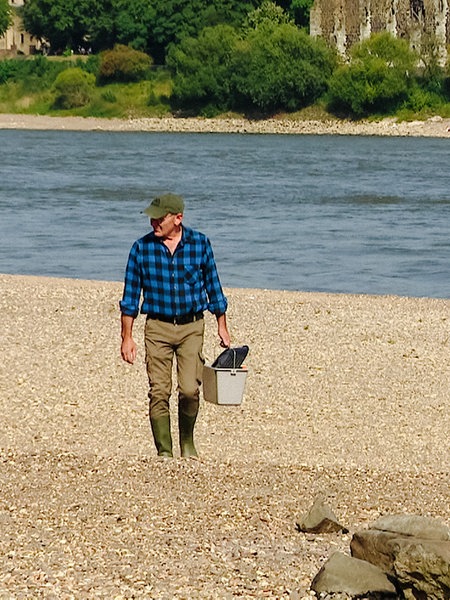 Jörg Ridder läuft auf einem Kieselstein-Strand entlang des Rheins. In der Hand hält er einen kleinen Eimer.