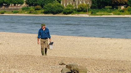Jörg Ridder läuft auf einem Kieselstein-Strand entlang des Rheins. In der Hand hält er einen kleinen Eimer.