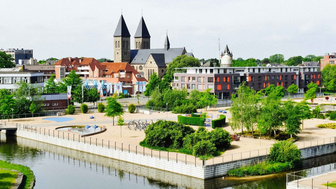 Der Inselpark Gronau mit einer Kirche und mehereren Häusern im Hintergrund. Auf dem Inselpark befinden sich Bäume, Wasserflächen und Spielgeräte für Kinder.