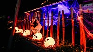 Es ist Nacht. Ein ungleichmäßiger Holzzaun steht vor einem Grusel-Haus. Davor liegen leuchtende Totenköpfe.